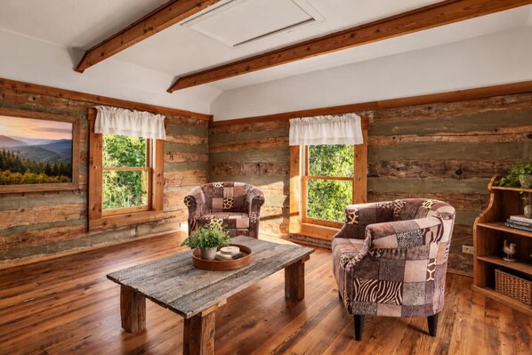 A rustic cabin interior with wooden walls, two upholstered chairs, a wooden table with a plant, and a bookshelf.