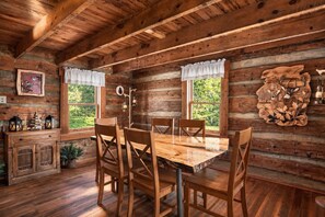 A rustic wooden cabin interior with a dining table, chairs, windows, and a fireplace.