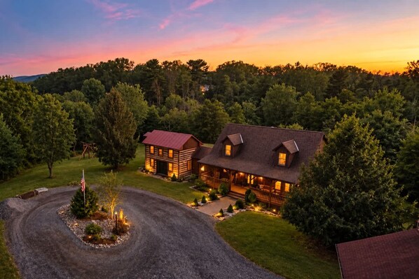 A two-story log cabin with a porch, surrounded by a forested landscape.