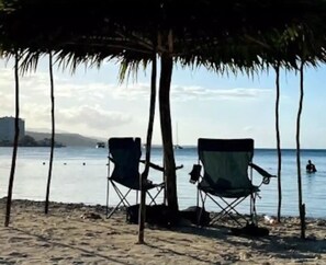 Ein Strand mit zwei Liegen unter einer Palme, ruhigem Meer und einer entfernten Küste mit Gebäuden.
