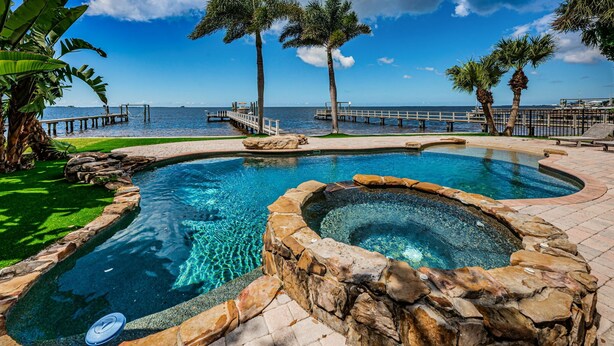A swimming pool with a hot tub, surrounded by palm trees and a dock extending into the water.