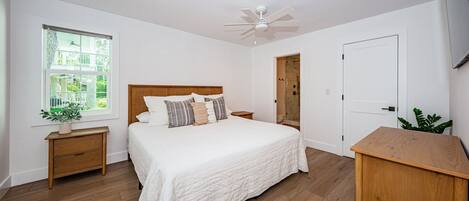 Un dormitorio con cama de madera, paredes blancas, ventilador de techo, ventana con vistas y mesita de noche de madera con una planta.