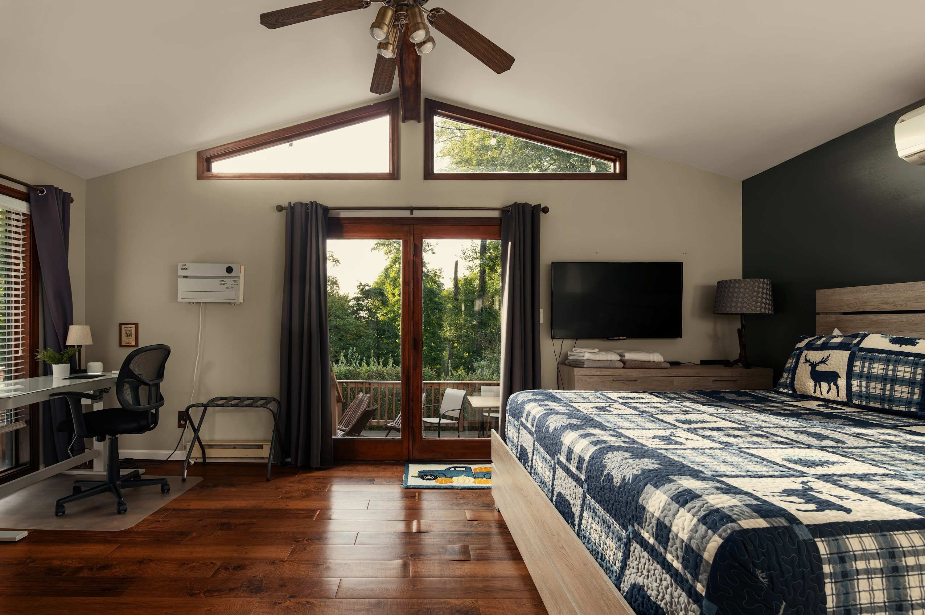 Un dormitorio con una cama grande, un escritorio con silla, un televisor y un balcón con vistas a árboles.