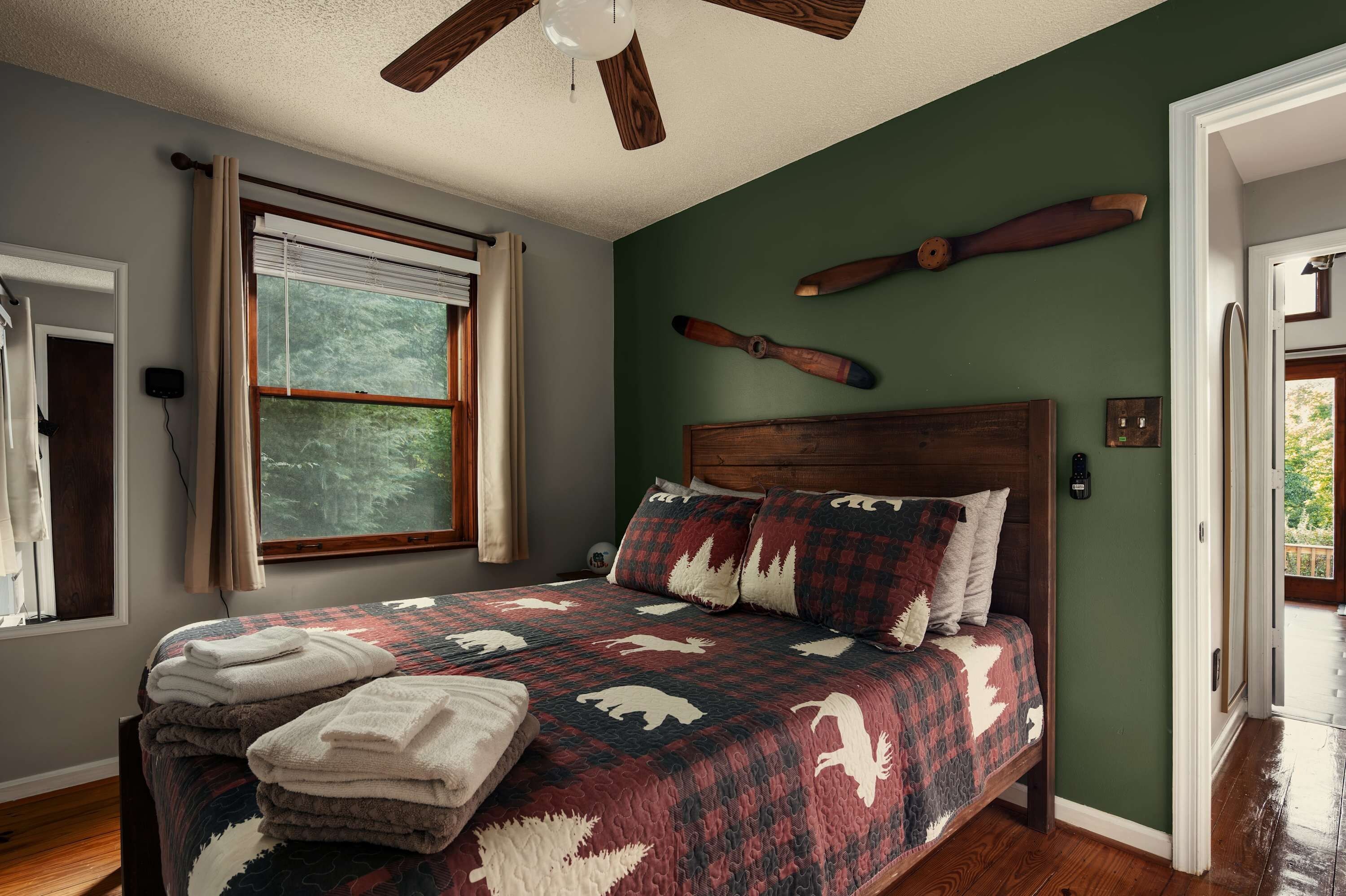 A bedroom with a wooden headboard, a large window with curtains, a ceiling fan, and a quilted bedspread.