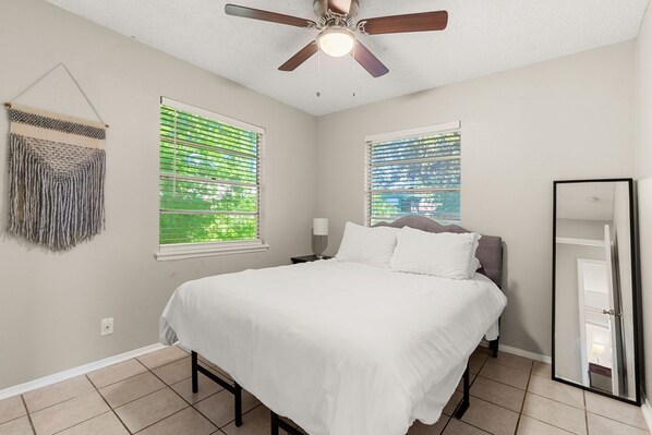 A bedroom with a bed, two windows with blinds, a ceiling fan, and a wall-mounted mirror.