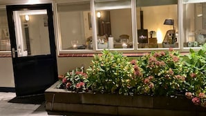 A building with a black door and windows with lights on inside. There is a planter with green plants and pink flowers in front of the building.