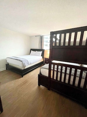 Quarto com duas camas, uma com encosto e outra sem, janela com cortinas e piso de madeira.