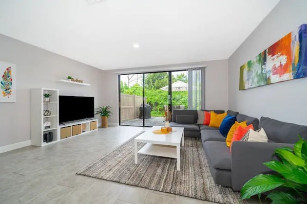 Relax in this stylish living room with a large sofa, flat-screen TV, and sliding glass door leading to a serene outdoor space.