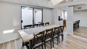A modern dining area with a long wooden table and dark chairs, white walls, and a large window.