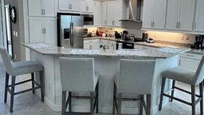 A modern kitchen with white cabinets, a central island with bar stools, stainless steel appliances, and a large window. - Stunning Executive Home in Gated Community (Fort Myers)