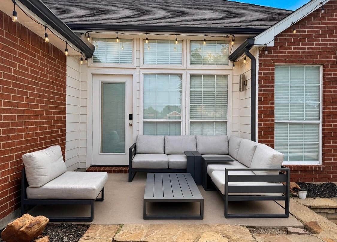 A covered patio with a sectional sofa, a coffee table, and a small table, all with metal frames and light-colored cushions.