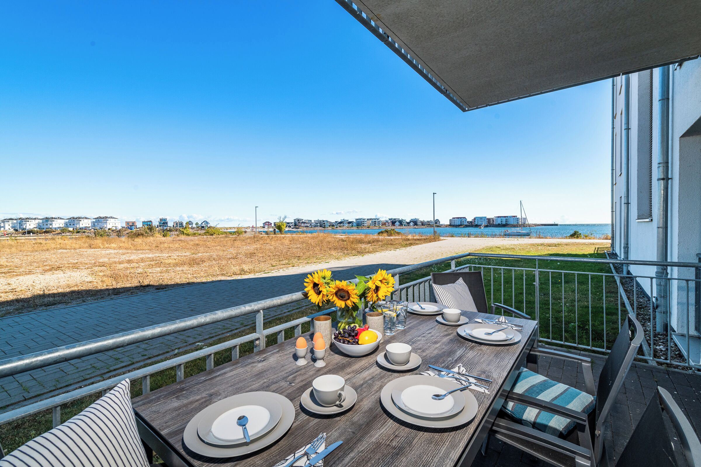 Une table à manger dressée avec des assiettes, des tasses et des couverts, sur un balcon donnant sur une plage et l’océan.