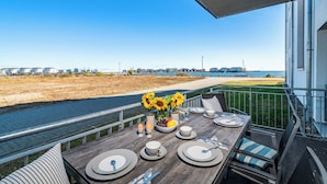 Mesa de comedor lista con platos, vasos y cubiertos en un balcón con vista a la playa y al agua.
