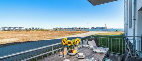 Une table à manger dressée avec des assiettes, des tasses et des couverts, sur un balcon donnant sur une plage et l’océan.