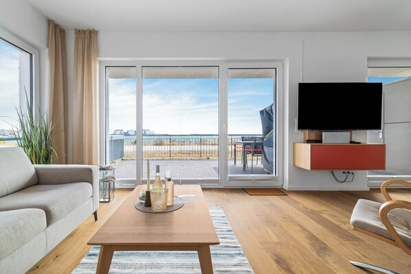 A modern living room with a sofa, a coffee table with bottles, a TV, and a sliding glass door to a balcony. - "Ocean Love": A vacation on the Baltic Sea, just a 3-minute walk from the beach, with a fireplace and a view of the sea (Olpenitz)