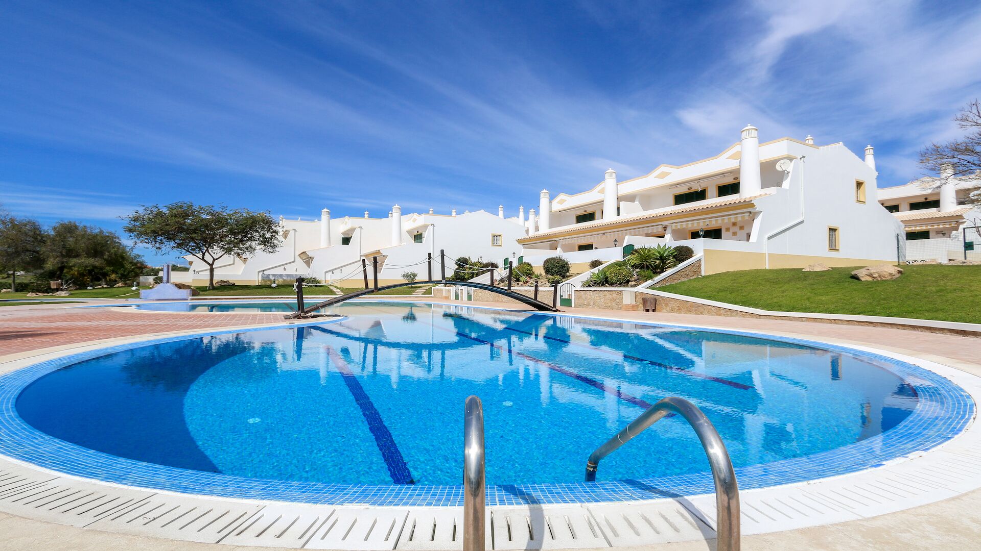 A swimming pool with a bridge, surrounded by white buildings with balconies, under a clear sky.
