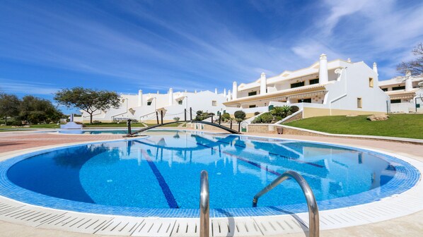 A swimming pool with a bridge, surrounded by white buildings with balconies, under a clear sky.