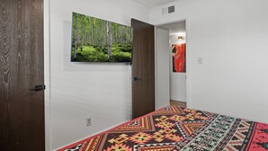 Une chambre avec une couvre-lit à motifs, un grand tableau sur le thème de la forêt accroché au mur et une vue sur une autre pièce.