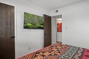 A bedroom with a patterned bedspread, a large forest-themed painting on the wall, and a view into another room. - Blushing Bungalow w/AC, Fenced Yard, W/D, Garage, Fast WiFi, 3 Samsung Smart TVs (Flagstaff)