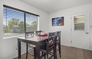 Un coin repas avec une table et des chaises, une fenêtre avec des stores, une porte avec un panneau de verre et un tableau encadré au mur.