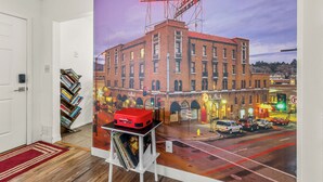 L’intérieur d’un hôtel, avec une grande fresque murale représentant un bâtiment sur lequel on peut lire « HÔTEL MONTE VISTA », une valise rouge posée sur une table et une bibliothèque remplie de livres.
