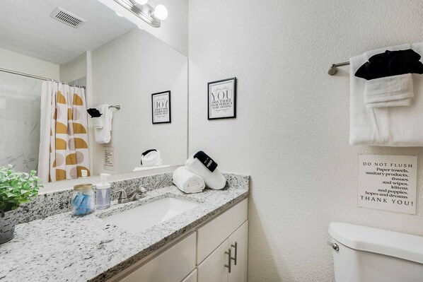 Une salle de bain moderne avec un comptoir en granit, un lavabo, un miroir et des toilettes. On y trouve des serviettes suspendues à un porte-serviettes, une citation encadrée accrochée au mur et un panneau rappelant de ne pas jeter les essuie-tout dans les toilettes.