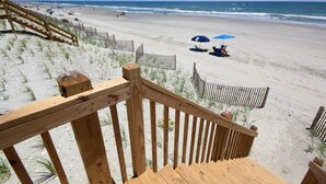 Un escalier en bois descend vers une plage de sable, bordée d’une clôture en bois et agrémentée d’un parasol bleu.