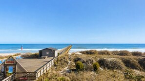 Une jetée en bois mène à un petit bâtiment surmonté d’un drapeau, qui surplombe une plage avec des dunes et l’océan.