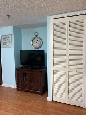 Une pièce comprenant un meuble en bois, un téléviseur, une horloge et une armoire blanche à portes à persiennes.