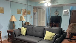 Una sala de estar con un sofá modular, un televisor, un ventilador de techo y un comedor con una mesa y sillas.