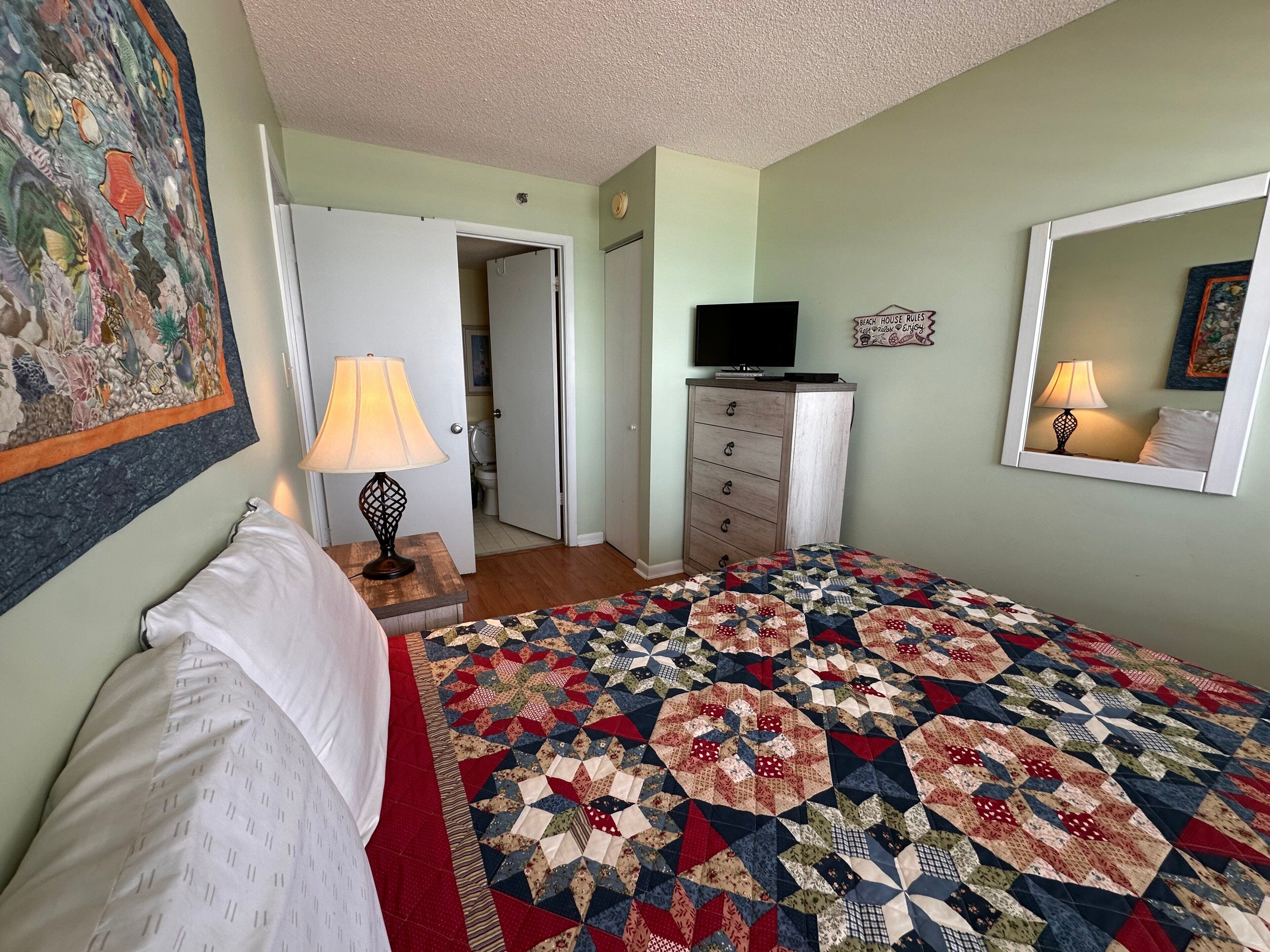 A bedroom with a bed, a dresser, a television, a lamp, and a door leading to a bathroom.