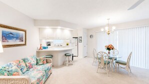 A modern kitchen with white cabinets, a dining area with a glass table and wicker chairs, and a living room with a floral sofa. - Closest Shorehaven Unit to Cherry Grove Beach • 4-Min Walk • Sleeps 6 (North Myrtle Beach)