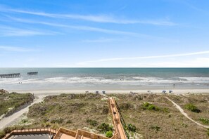 A beach view with a wooden boardwalk leading to the water, a pier in the distance, and a clear sky.