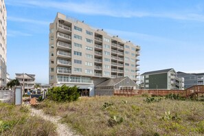 A multi-story residential building with balconies, a covered entrance, and a sandy foreground with vegetation.