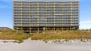 Un hôtel à plusieurs étages, doté de balcons, situé sur une plage de sable fin, avec une végétation luxuriante en premier plan.