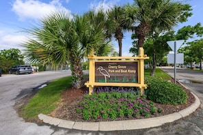 Willkommensschild für den Cherry Grove Park und Bootsrampe, mit einer Vogelillustration, umgeben von Palmen und violetten Blumen.