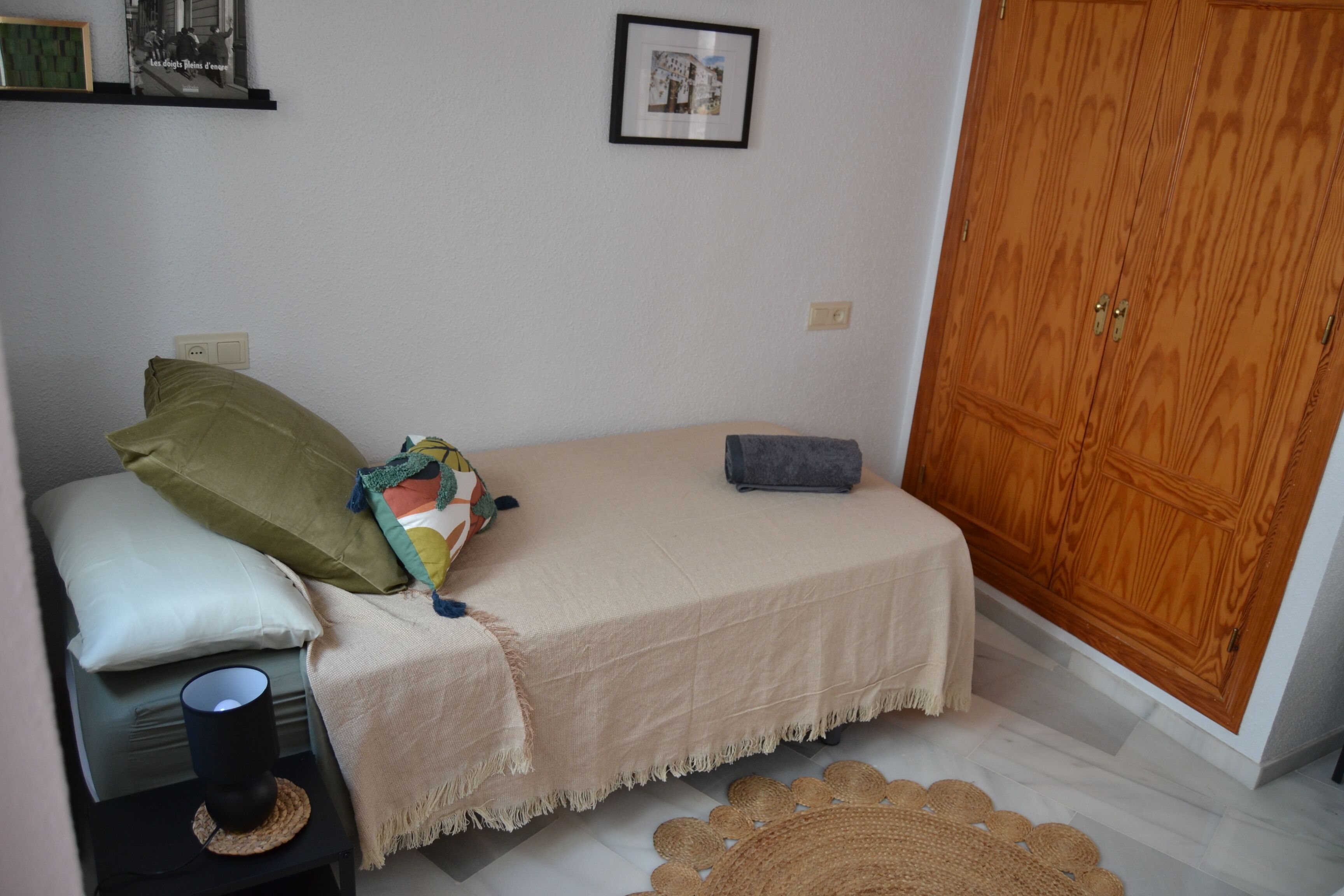 A small bedroom with a bed, pillows, a bedside table with a lamp, a wooden wardrobe, and a woven mat on the floor.