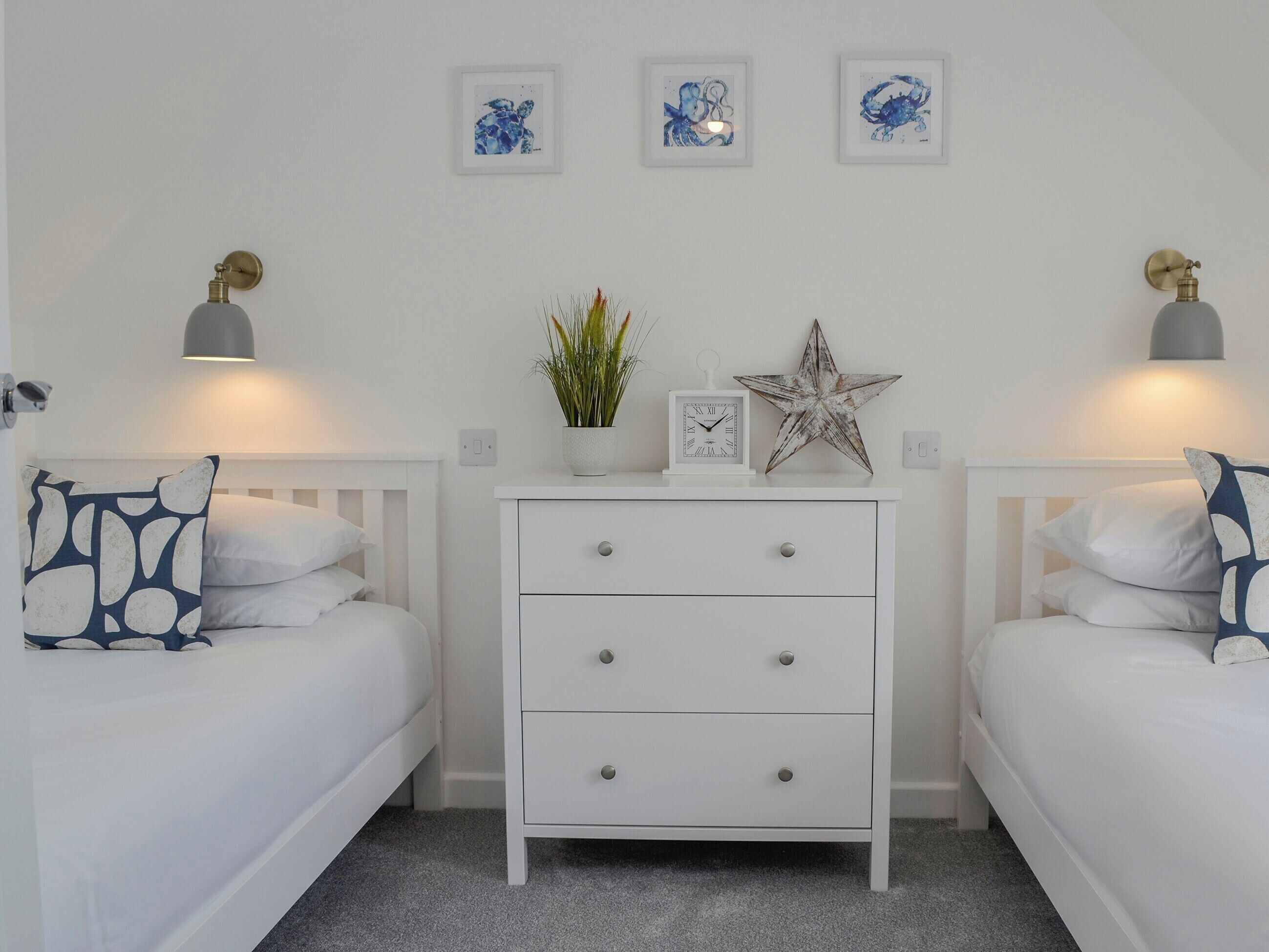 Un dormitorio con dos camas blancas, una mesita de noche blanca con cajones, un reloj de pared, una decoración con forma de estrella y cuadros en la pared sobre la mesita de noche.
