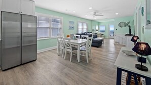 A spacious kitchen with a dining area, a living room with a sofa and a coffee table, and a view of the ocean through the windows. - Oceanfront 5BR Home • Back Deck on the Beach • Cherry Grove • Momma’s Gift (North Myrtle Beach)
