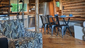 Interior rústico de una cabaña con techo de madera, una zona de comedor con sillas y una mesa, un sofá y un refrigerador.