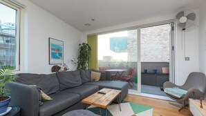 Ein modernes Wohnzimmer mit einem grauen Sofa, einem kleinen Holztisch und einem Essbereich mit Tisch und Stühlen. Ein Glastür öffnet sich zu einem Balkon mit Blumentöpfen.