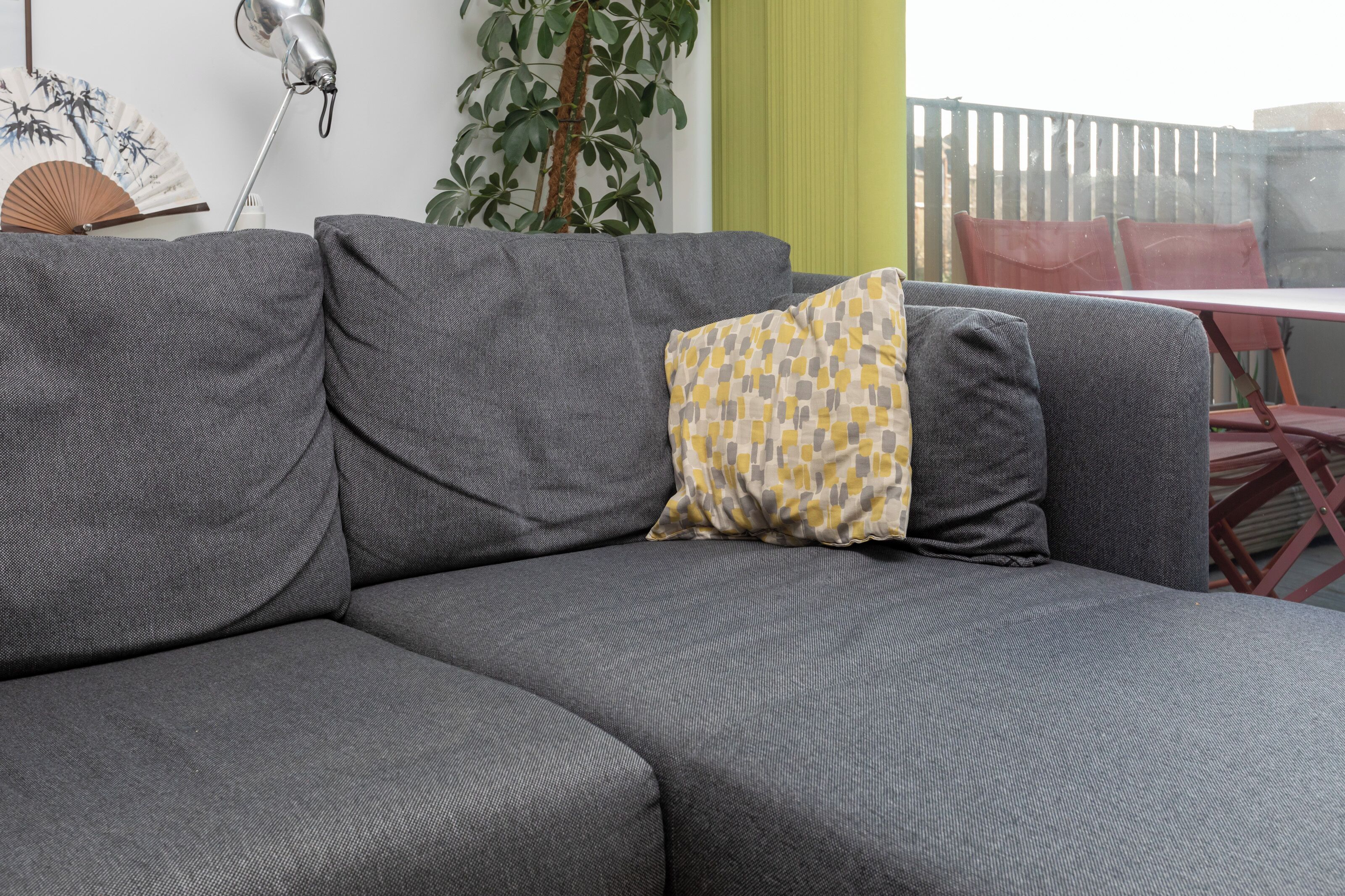 Un canapé gris agrémenté d’un coussin à motifs, une lampe fixée au mur, une plante et une vue sur un patio extérieur avec une table et des chaises.