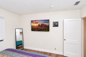 Ein Zimmer mit einem Bett, einem Wandspiegel, einer Tür und einem Bilderrahmen mit einer Sonnenlandschaft.