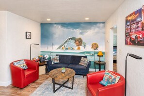 Ein Wohnzimmer mit einem blauen Sofa, roten Sesseln, einem Couchtisch und einem Wandbild, das ein Auto und den Himmel zeigt.