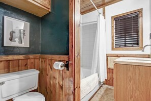 Un baño con tocador de madera, inodoro, ducha con cortina y ventana con persianas.