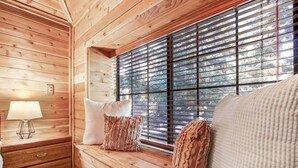 Interior acollidor d'una cabana, amb una banca de fusta, coixins, una làmpada i una finestra amb persianes.