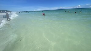 Een strandscène met helder turquoise water, een zandstrand en enkele mensen die zwemmen en wandelen.