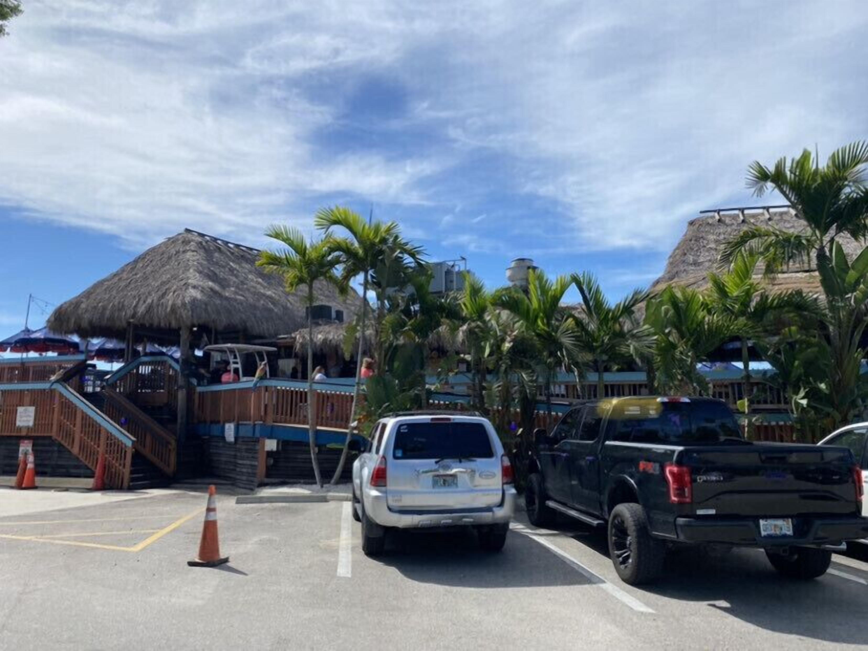 A parking area with a white SUV and a black pickup truck. There are palm trees and a thatched-roof structure in the background.