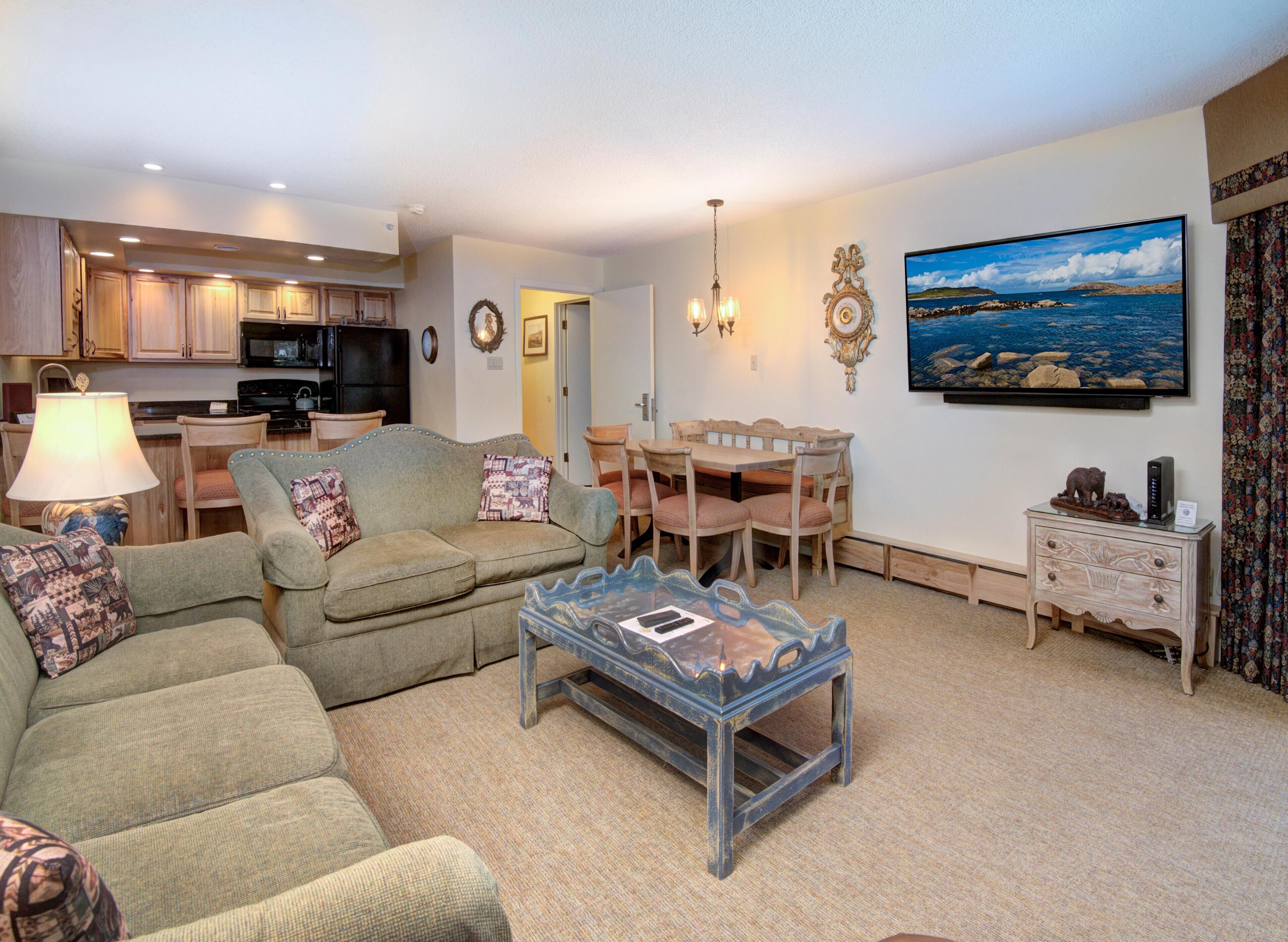 A living room with a sofa, a coffee table, a dining table with chairs, a kitchen area, and a painting on the wall.