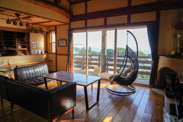 A cozy room with a wooden floor, a black leather sofa, a small table, and a hanging chair. A large window offers a view of a wooden deck and greenery. - 5LDK with natural hot spring and the ocean view - Gift Villa / Beppu Ōita (Beppu)